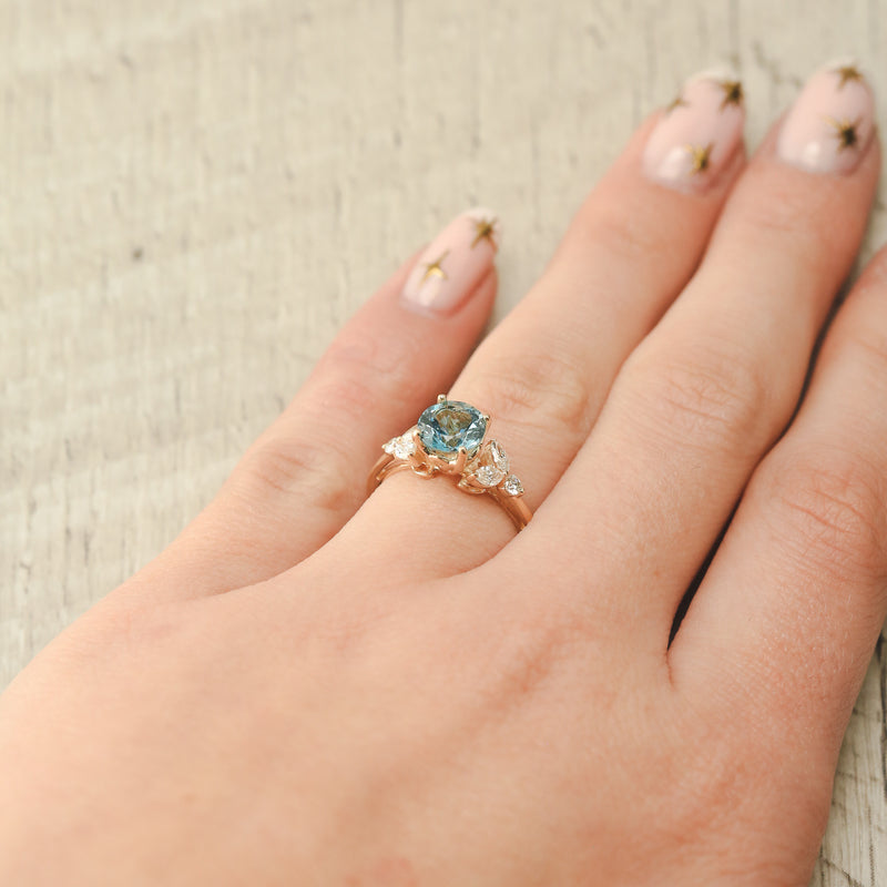 Shown here, Blossom, is a round cut aquamarine women's engagement ring with leaf-shaped diamond accents on hand. Many other center stone options are available upon request. 