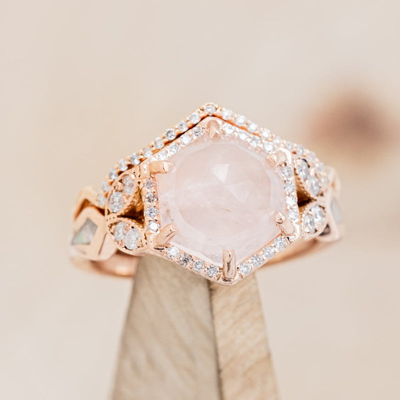  Shown here is "Lucy in the Sky" is a halo-style hexagon faceted rose quartz women's engagement ring with diamond accents, mother of pearl inlays, and a diamond tracer, on stand tilted left. Many other center stone options are available upon request. 