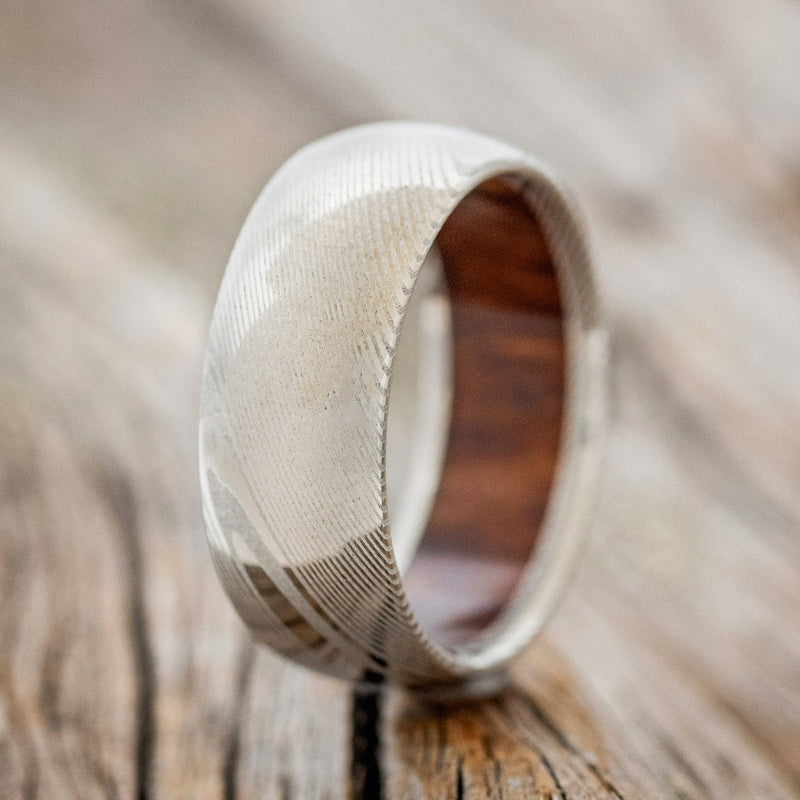 Shown here is a handcrafted men's wedding ring with an ironwood lining on a domed Damascus steel band, upright facing left. Additional inlay options are available upon request.