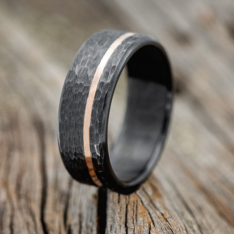 Shown here is a handcrafted men's wedding ring featuring an offset thin 14k rose gold inlay and a hammered finish, upright facing left. Shown here set on a fire treated black zirconium wedding band. Additional inlay options are available upon request.