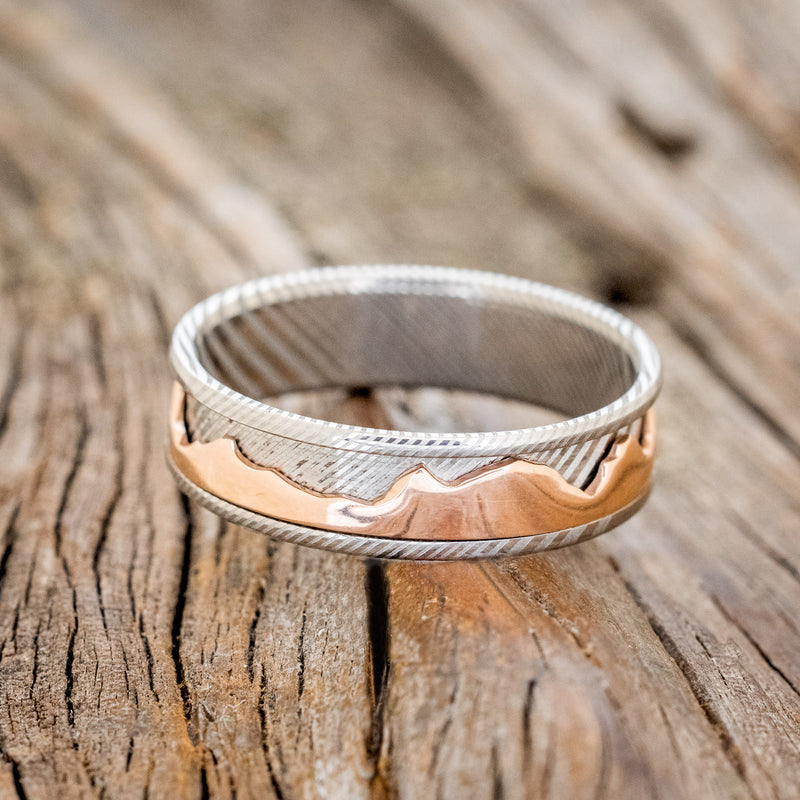 Shown here is "Revolution", a handcrafted fidget men's wedding ring featuring a free-spinning 14K rose gold mountain range on a Damascus steel band, laying flat. Additional inlay options are available upon request.
