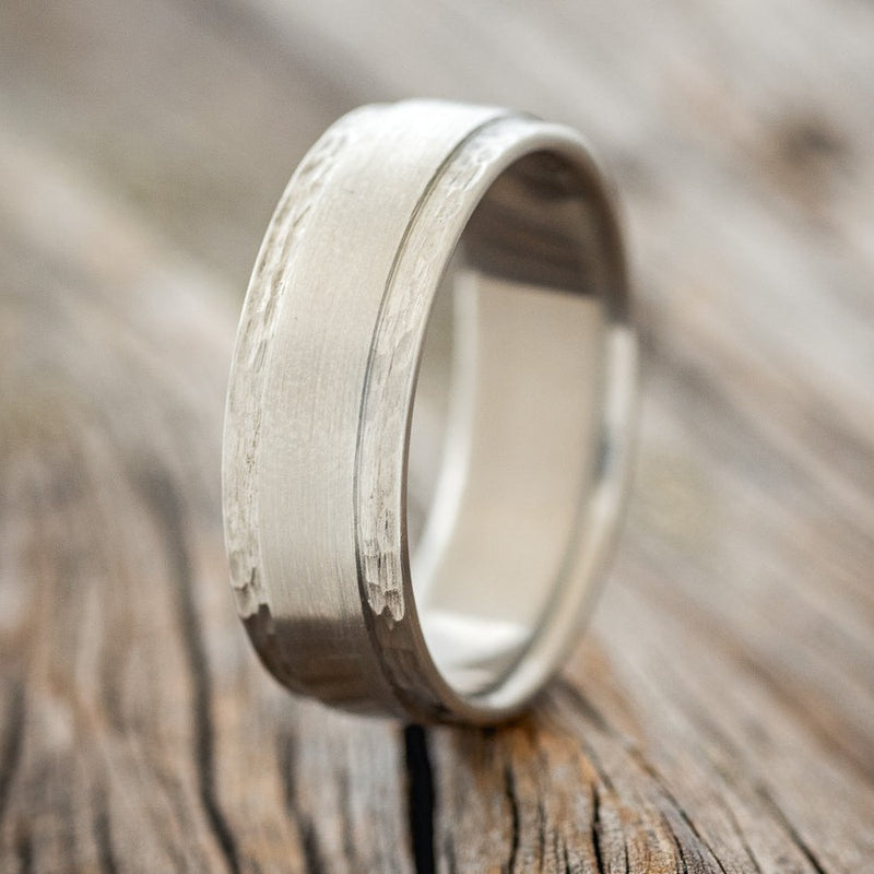 Shown here is "Sedona", a custom, handcrafted men's wedding ring featuring a raised center with a brushed finish and hammered edges on a solid metal band, upright facing left. Additional inlay options are available upon request.