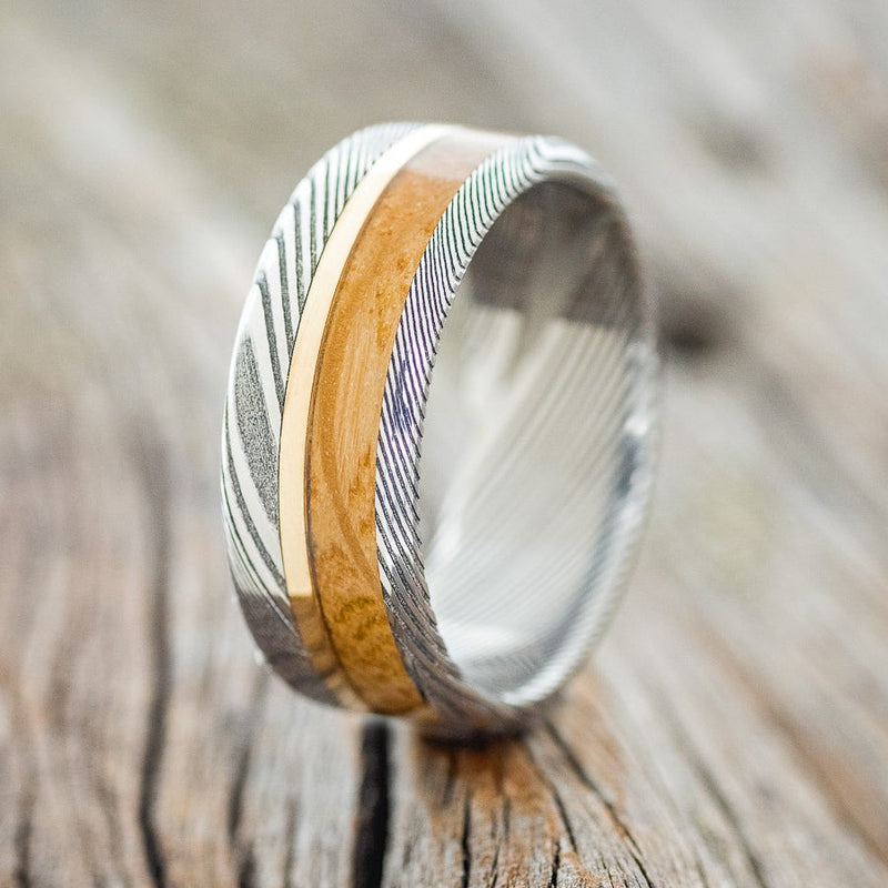 Shown here is "Tanner", a custom, handcrafted men's wedding ring featuring a whiskey barrel overlay and a 14K yellow gold inlay, shown here on a Damascus steel band, upright facing left. Additional inlay options are available upon request.