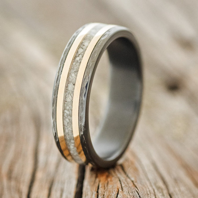 Shown here is "Hollis", a custom, handcrafted men's wedding ring featuring a hammered, fire-treated black zirconium band with elk tooth ivory & two 14K yellow gold inlays, upright facing left. Additional inlay options are available upon request.