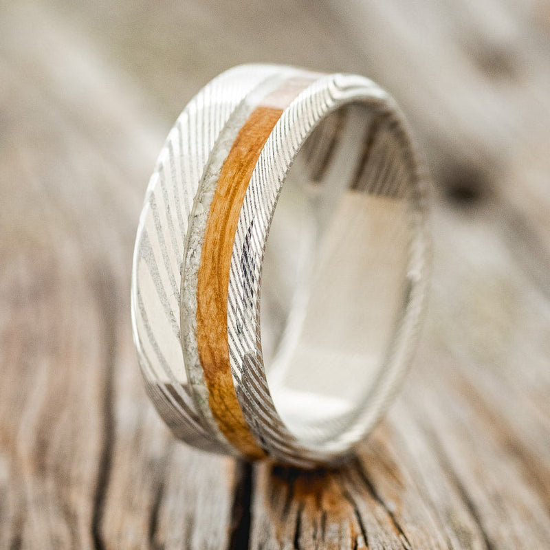 Shown here is "Castor", a custom, handcrafted men's wedding ring featuring an elk tooth and whiskey barrel oak inlay on a Damascus steel band, upright facing left. Additional inlay options are available upon request.