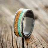 Shown here is "Dyad", a custom, handcrafted men's wedding ring featuring 2 channels with fire and ice opal, malachite, and whiskey barrel oak inlays, upright facing left.