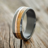 Shown here is "Tanner", a custom, handcrafted men's wedding ring featuring a whiskey barrel and a 14K rose gold inlay, shown here on a fire-treated black zirconium band, upright facing left. Additional inlay options are available upon request.