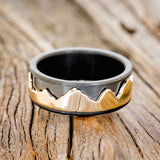 Shown here is "Revolution", a handcrafted fidget men's wedding ring featuring a free-spinning 14K yellow gold mountain range on a fire-treated black zirconium band, laying flat. Additional inlay options are available upon request.