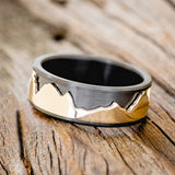 Shown here is "Revolution", a handcrafted fidget men's wedding ring featuring a free-spinning 14K yellow gold mountain range on a fire-treated black zirconium band, tilted left. Additional inlay options are available upon request.