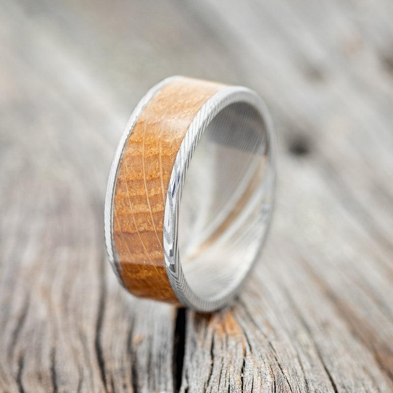 Shown here is "Rainier", a custom, handcrafted men's wedding ring featuring whiskey barrel wood inlay on a Damascus steel band, upright facing left. Additional inlay options are available upon request.