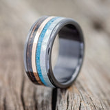 Shown here is a custom, handcrafted men's wedding ring featuring a single channels with mother of pearl, turquoise, 14K rose gold inlay and patina copper, shown here on a fire-treated black zirconium band, upright facing left. Additional inlay options are available upon request.