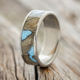 Shown here is "Rainier", a custom, handcrafted men's wedding ring featuring buckeye burl wood with hand-crushed turquoise filling the knot sand burl holes on a Damascus Steel band, upright facing left. Additional inlay options are available upon request.