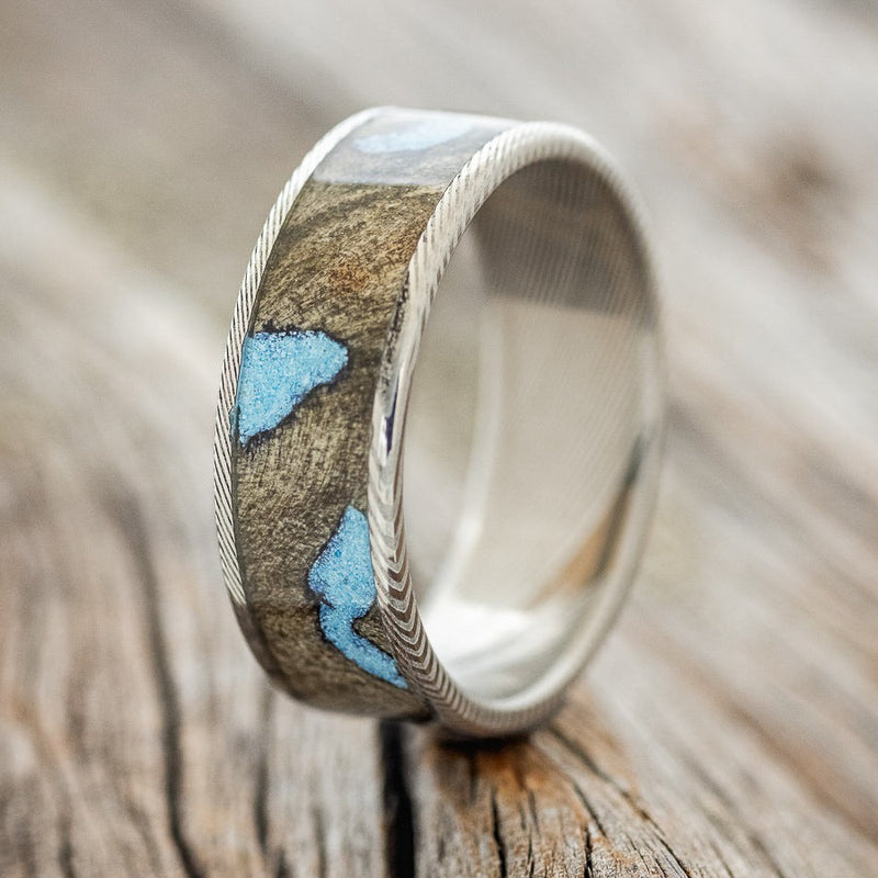 Shown here is "Rainier", a custom, handcrafted men's wedding ring featuring buckeye burl wood with hand-crushed turquoise filling the knot sand burl holes on a Damascus Steel band, upright facing left. Additional inlay options are available upon request.