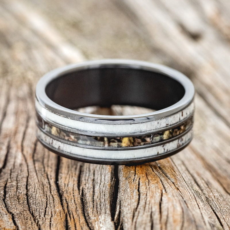 Shown here is "Rio", a custom, handcrafted men's wedding ring featuring 3 channels with antler and camo inlays on a fire-treated black zirconium band, laying flat. Additional inlay options are available upon request.