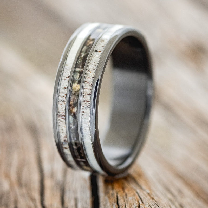 Shown here is "Rio", a custom, handcrafted men's wedding ring featuring 3 channels with antler and camo inlays on a fire-treated black zirconium band, upright facing left. Additional inlay options are available upon request.