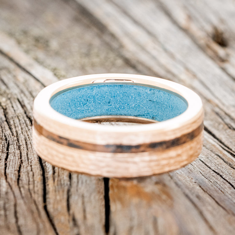 Shown here is "Vertigo", a custom, handcrafted men's wedding ring featuring an offset copper patina inlay and a turquoise lining on a hammered 14K gold band, laying flat. Additional inlay options are available upon request.