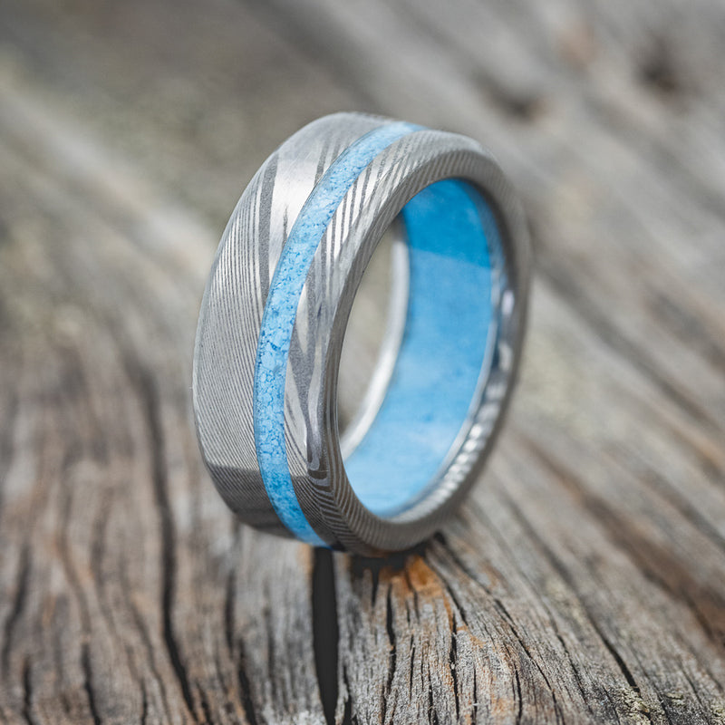 Shown here is "Vertigo", a handcrafted men's wedding ring featuring a turquoise lining and an offset turquoise inlay shown on a Damascus steel wedding band, upright facing left. Additional inlay options are available upon request.