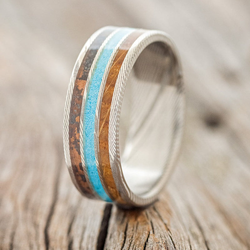 Shown here is "Rio", a custom, handcrafted men's wedding ring featuring 3-channels of patina copper, turquoise, and whiskey barrel oak on a Damascus steel band, upright facing left. Additional inlay options are available upon request.