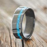Shown her is "Vertigo", a custom, handcrafted men's wedding ring featuring a turquoise inlay with a diagonal fluted on a fire-treated black zirconium band, upright facing left. Additional inlay options are available upon request.
