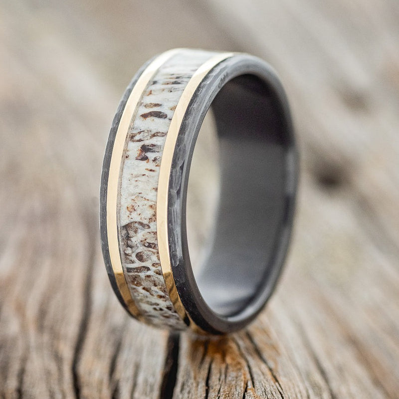 Shown here is "Hollis", a custom, handcrafted men's wedding ring featuring elk antler & 2 14K yellow gold inlays (1mm) on hammered, fire-treated black zirconium band, upright facing left. Additional inlay options are available upon request.