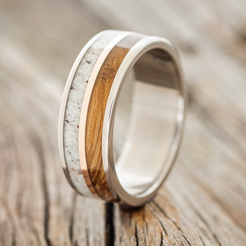 Shown here is "Dyad", a custom, handcrafted men's wedding ring featuring 2 channels with whiskey barrel and antler divided by a 14K rose gold inlay, shown here on a titanium band, upright facing left. Additional inlay options are available upon request.