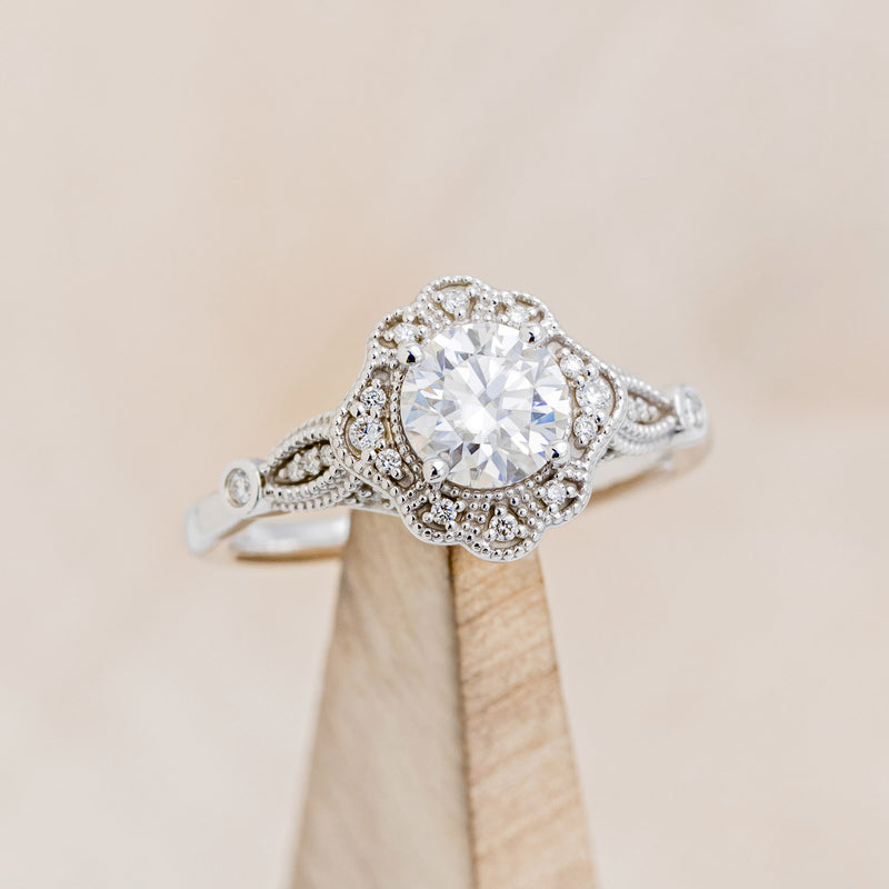 Shown here is "Eileen", a vintage-style moissanite women's engagement ring with a diamond halo, accents, on stand facing slightly right. Many other center stone options are available upon request.