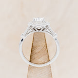 Shown here is "Eileen", a vintage-style moissanite women's engagement ring with a diamond halo, accents, side view on stand. Many other center stone options are available upon request.