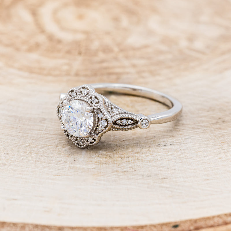 Shown here is "Eileen", a vintage-style moissanite women's engagement ring with a diamond halo, accents, facing left. Many other center stone options are available upon request.