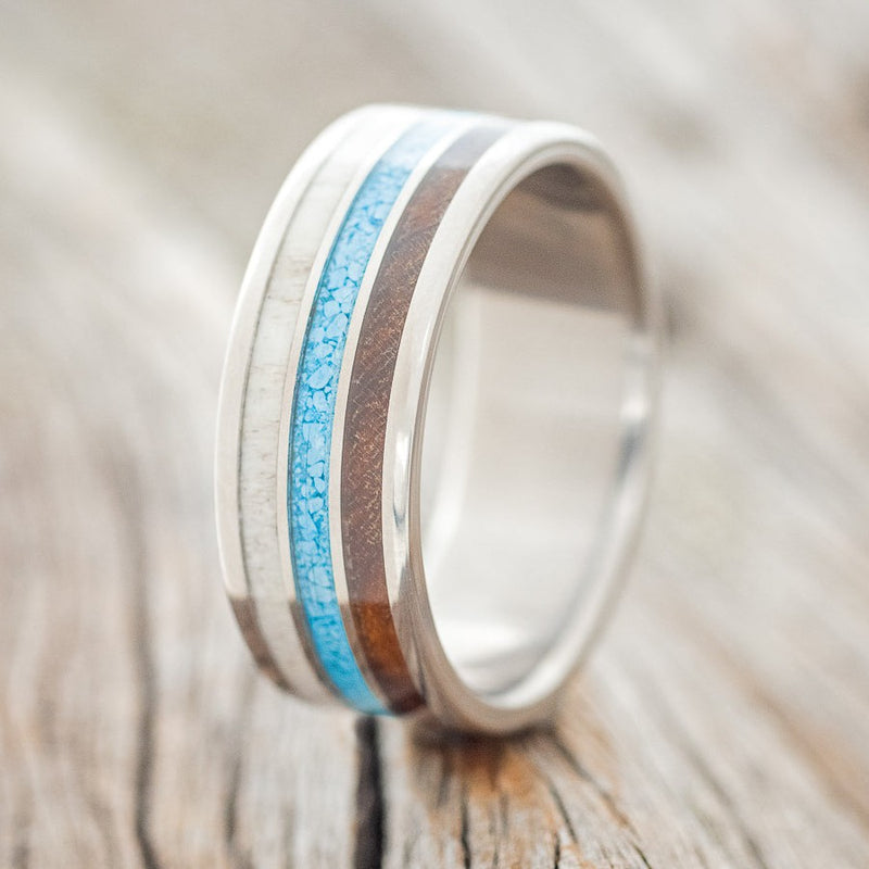 Shown here is "Rio", a custom, handcrafted men's wedding ring featuring 3 channels with elk antler, turquoise, and ironwood inlays on a titanium band, upright facing left. Additional inlay options are available upon request.