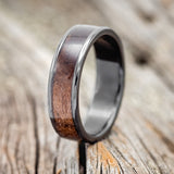 Shown here is "Rainier", a custom, handcrafted men's wedding ring featuring a dark maple wood inlay on a fire-treated black zirconium band, upright facing left. Additional inlay options are available upon request.