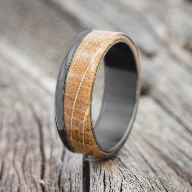 Shown here is "Ezra", a custom, handcrafted men's wedding ring featuring a whiskey barrel oak overlay, shown here on a fire-treated black zirconium band, upright facing left. Additional inlay options are available upon request.