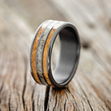 Shown here is "Rio", a custom, handcrafted men's wedding ring featuring 3 channels with a whiskey barrel and crushed black pearl inlays on a fire-treated black zirconium band, upright facing left. Additional inlay options are available upon request.
