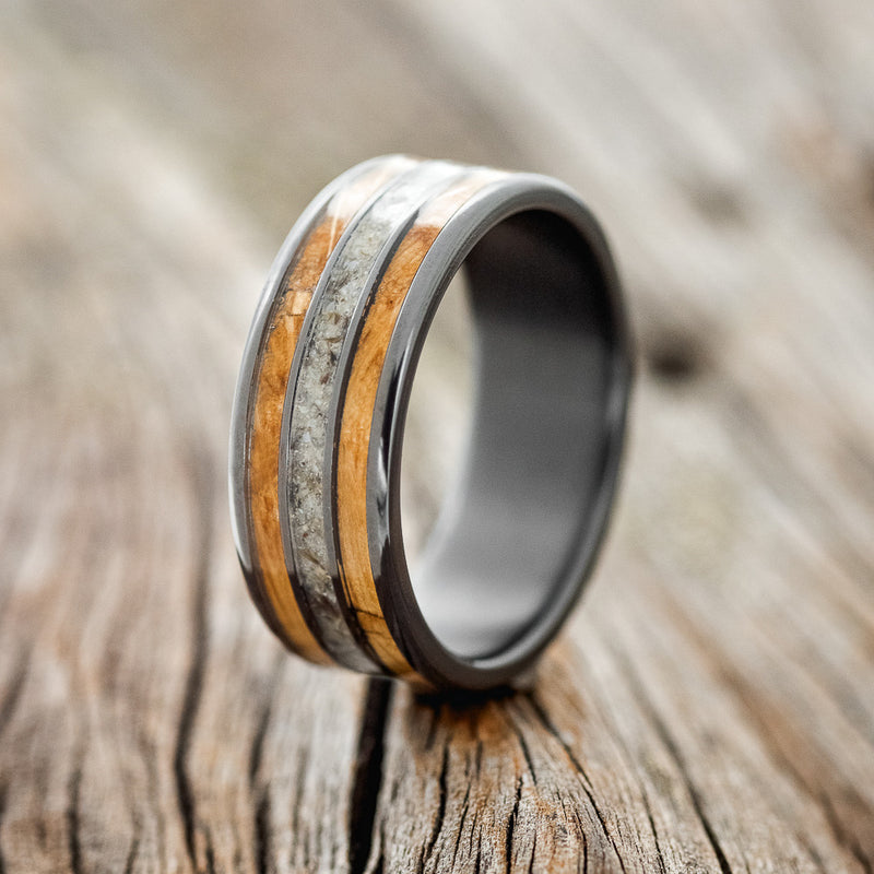 Shown here is "Rio", a custom, handcrafted men's wedding ring featuring 3 channels with a whiskey barrel and crushed black pearl inlays on a fire-treated black zirconium band, upright facing left. Additional inlay options are available upon request.