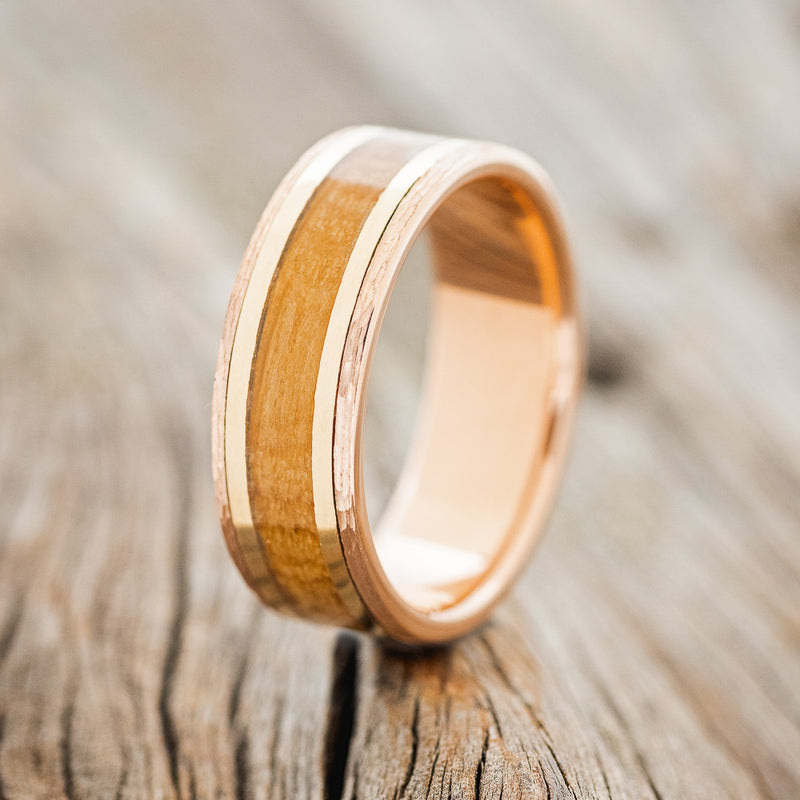 Shown here is "Hollis", a handcrafted men's wedding ring featuring a whiskey barrel oak & 2 14K yellow gold inlays (1mm) on a hammered, 14k gold band, upright facing left. Additional inlay options are available upon request.