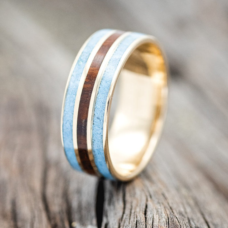 Shown here is "Rio", a custom, handcrafted men's wedding ring featuring 3 channels with turquoise and ironwood inlays on a 14K gold band, upright facing left. Additional inlay options are available upon request.