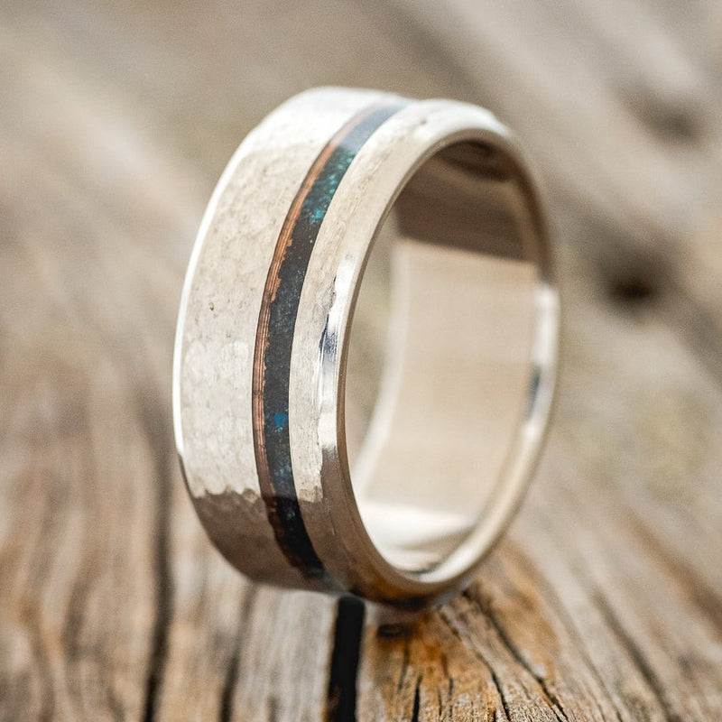 Shown here is "Vertigo", a custom, handcrafted men's wedding ring featuring patina copper inlay on a hammered titanium band, upright facing left. Additional inlay options are available upon request.