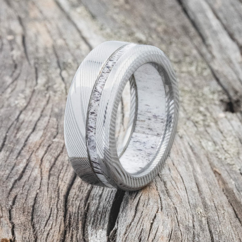 Shown here is "Vertigo", a handcrafted antler lined men's wedding ring featured a Damascus steel band with an offset antler inlay, upright facing left. All of our antler is ethically collected naturally. Additional wood and inlay options are available upon request.