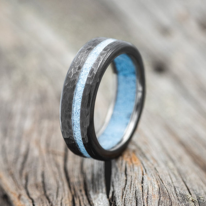 Shown here is "Vertigo", a custom, handcrafted men's wedding ring featuring a turquoise lining and inlay on a fire-treated, hammered black zirconium band, upright facing left. Additional inlay options are available upon request.