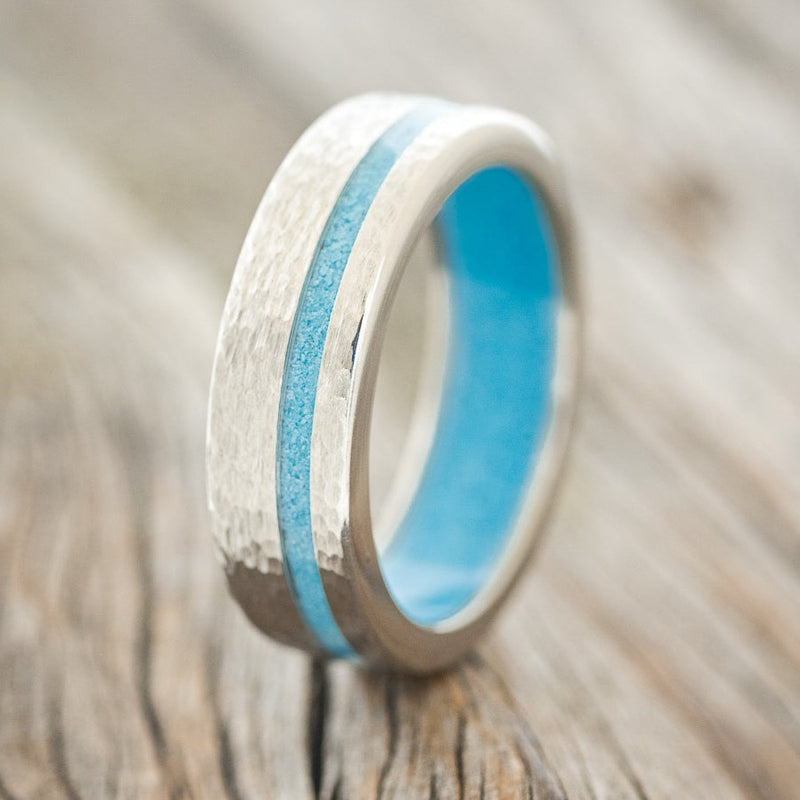 Shown here is "Vertigo", a custom, handcrafted men's wedding ring featuring a turquoise lining and inlay on a hammered titanium band, upright facing left. Additional inlay and lining options are available upon request.