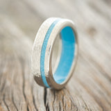 Shown here is "Vertigo", a custom, handcrafted men's wedding ring featuring a turquoise lining and inlay on a hammered band, upright facing left. 
