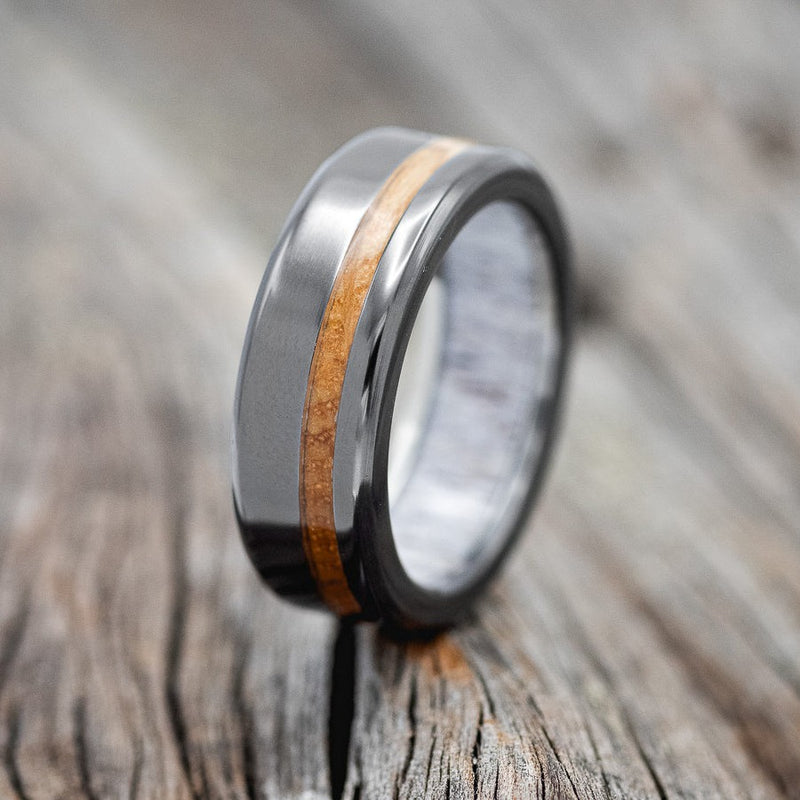 Shown here is "Vertigo", a handcrafted men's wedding ring featuring whiskey barrel inlay on fire-treated black zirconium band, with an elk antler lining, upright facing left. Additional inlay options are available upon request.
