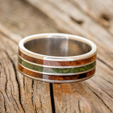 Shown here is "Rio", a custom, handcrafted men's wedding ring featuring 3 channels with redwood and moss inlays on a titanium band, laying flat. Additional inlay options are available upon request.