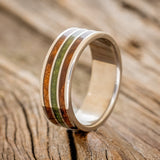 Shown here is "Rio", a custom, handcrafted men's wedding ring featuring 3 channels with redwood and moss inlays on a titanium band, upright facing left. Additional inlay options are available upon request.