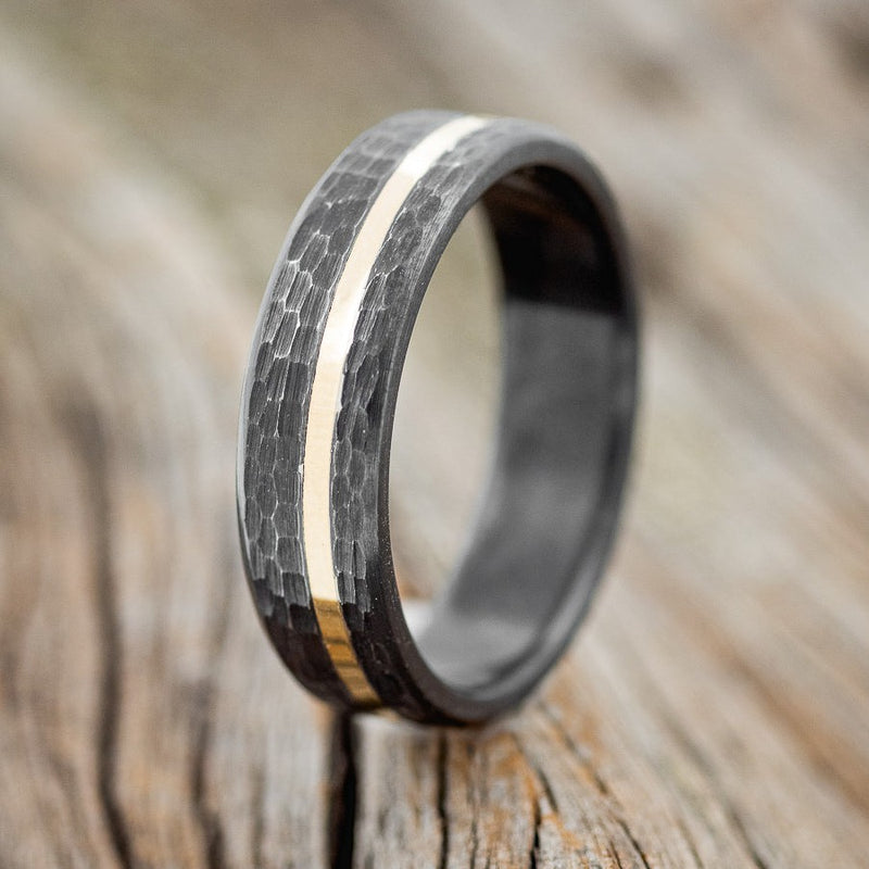 Shown here is a handcrafted men's wedding ring featuring an offset 14K yellow gold inlay with a hammered finish, upright facing left. Shown here set on a fire-treated black zirconium wedding band. Additional inlay options are available upon request.