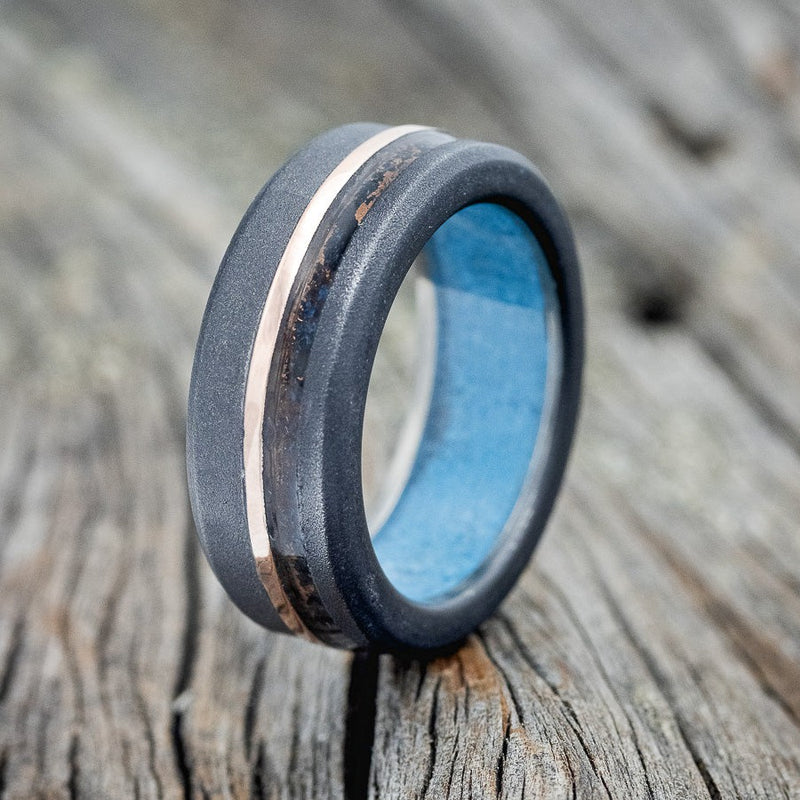Shown here is "Castor", a handcrafted men's wedding ring featuring a patina copper and 14K rose gold inlay on a sandblasted black zirconium band with a turquoise lining, upright facing left. Additional inlay options are available upon request.