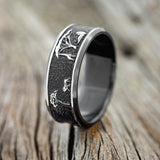 Shown here is "Legacy", a handcrafted, custom embossed men's wedding ring featuring an African savannah engraving in a channel-style band, upright facing left. This ring is pictured in black zirconium, giving it a higher contrast between embossment and engraving than other metal types will provide. It can be customized to feature just about any embossed design you can dream up.