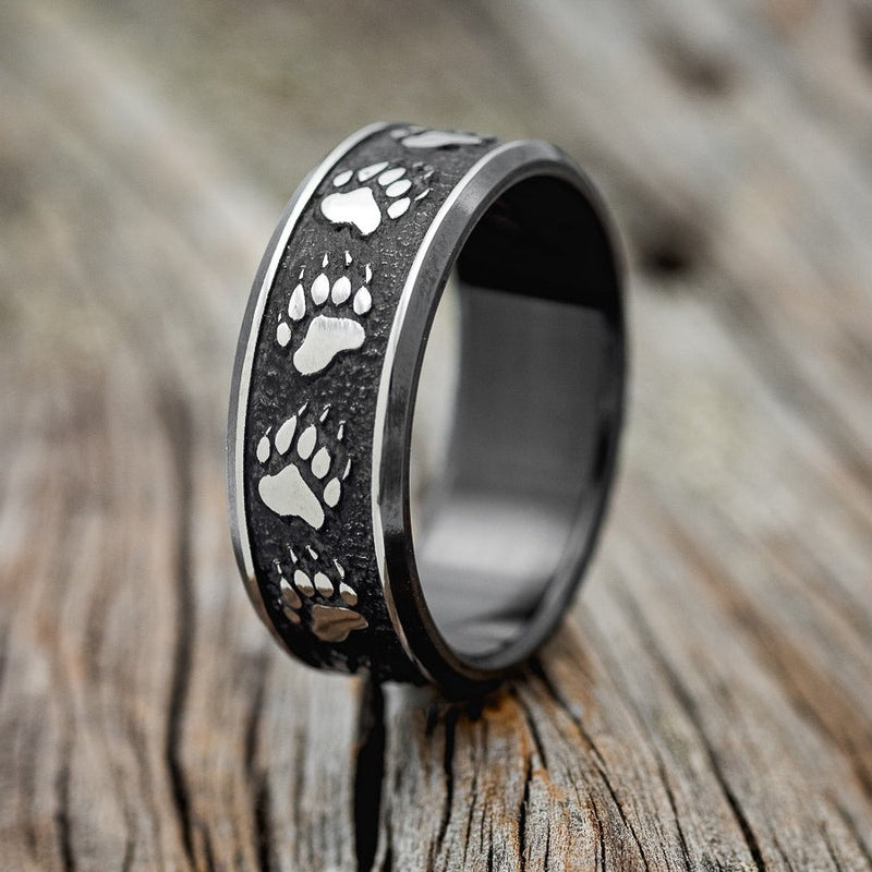 Shown here is "Legacy", a handcrafted, custom embossed men's wedding ring featuring a bear paw engraving in a channel-style band, upright facing left. This ring is pictured in black zirconium, giving it a higher contrast between embossment and engraving than other metal types will provide. It can be customized to feature just about any embossed design you can dream up.