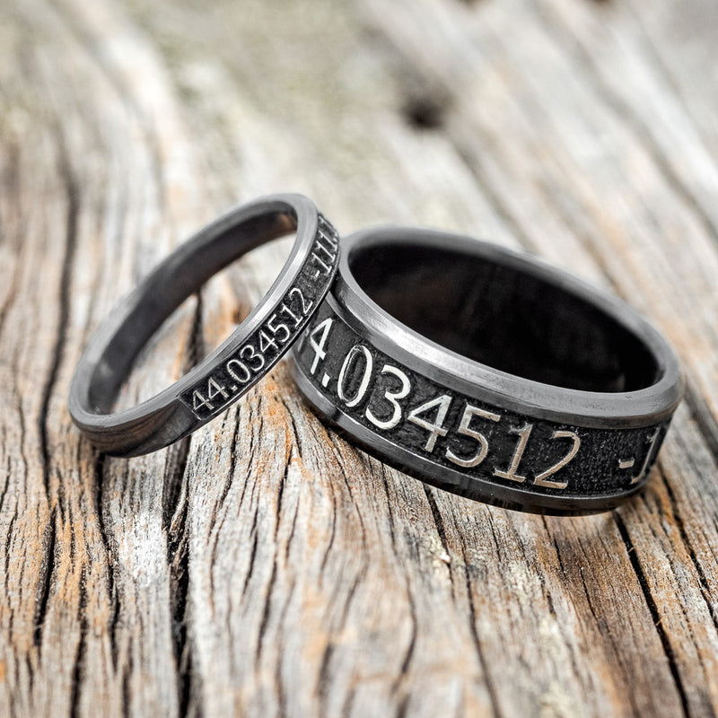 Shown here is "Path Finder", a handcrafted, matching custom embossed coordinates wedding bands, upright facing left. This set is pictured in black zirconium, giving it a higher contrast between embossment and engraving than other metal types will provide. 