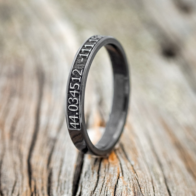 Shown here is "Path Finder", a handcrafted, custom embossed coordinates women's wedding band, upright facing left. This ring is pictured in black zirconium, giving it a higher contrast between embossment and engraving than other metal types will provide. It can be customized to feature just about any embossed design you can dream up.