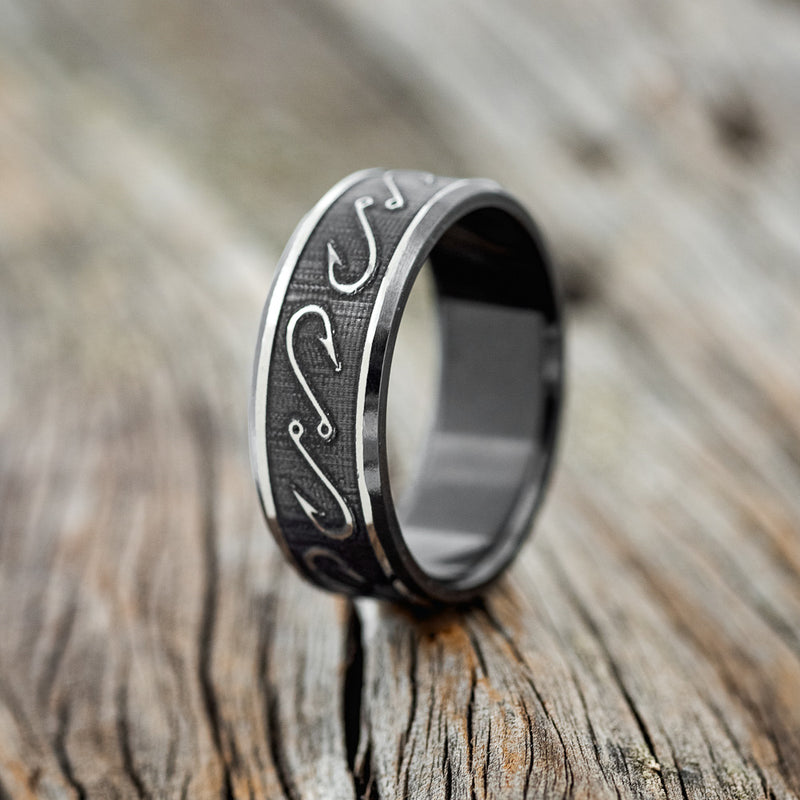 Shown here is "Legacy", a handcrafted, custom embossed men's wedding ring featuring a fishing hook engraving in a channel-style band, upright facing left. This ring is pictured in black zirconium, giving it a higher contrast between embossment and engraving than other metal types will provide. It can be customized to feature just about any embossed design you can dream up.